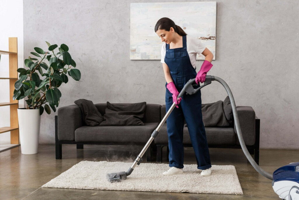 Beautiful cleaner in workwear and rubber gloves using vacuum cleaner with hot steam on carpet in